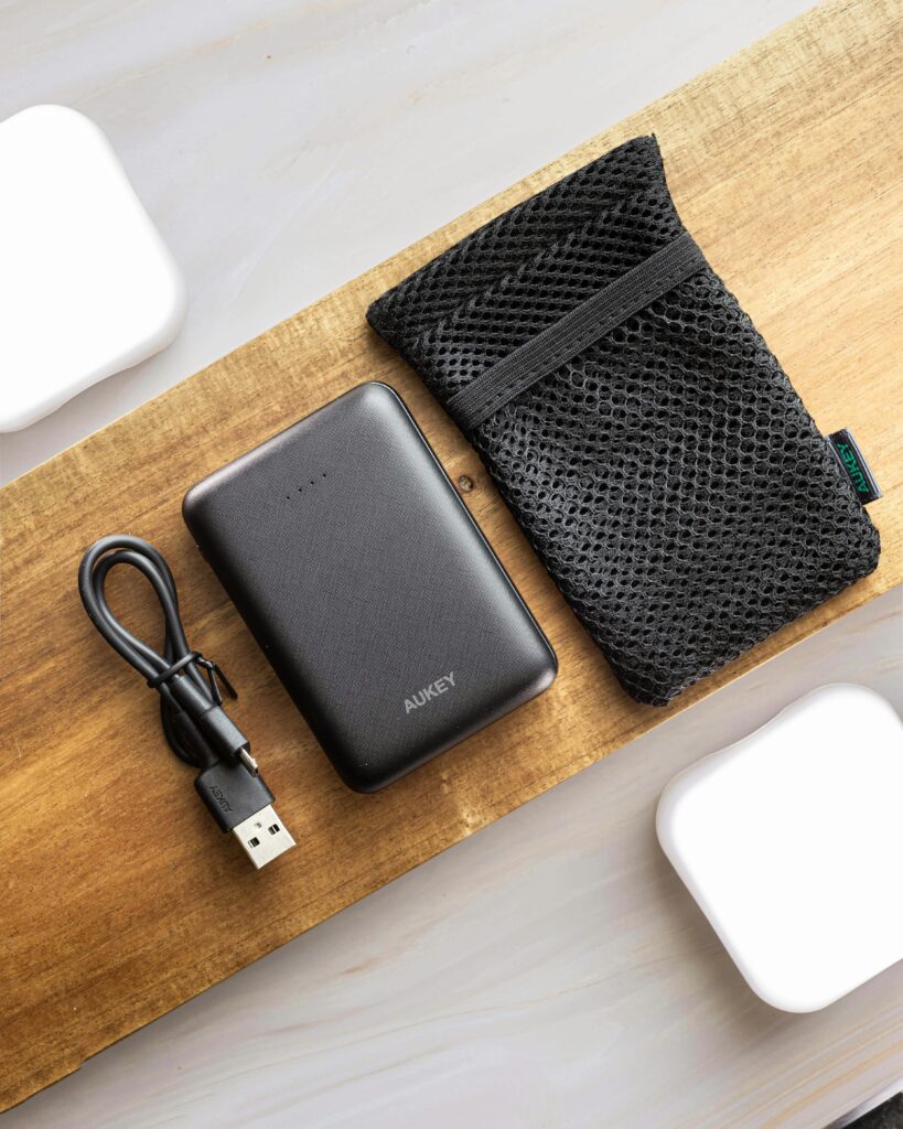 Flat lay of a black power bank with a USB cable and mesh pouch on wood.